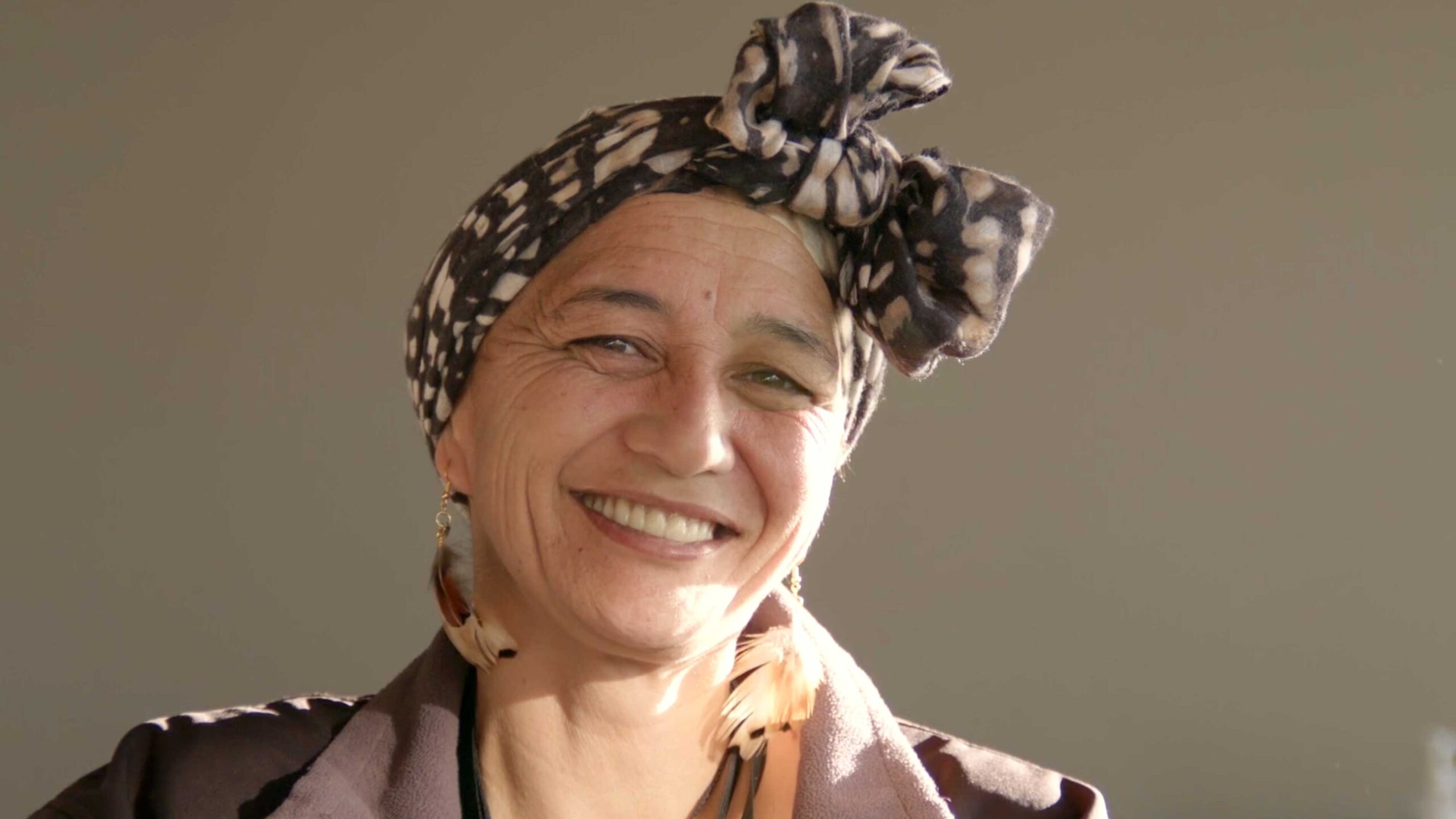 Woman smiling warmly, wearing a patterned head wrap and feather earrings