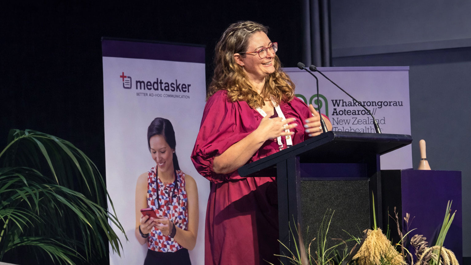 A woman in a red dress speaks at a podium at the Whakarongorau Aotearoa New Zealand Telehealth conference, with a Medtasker banner behind her
