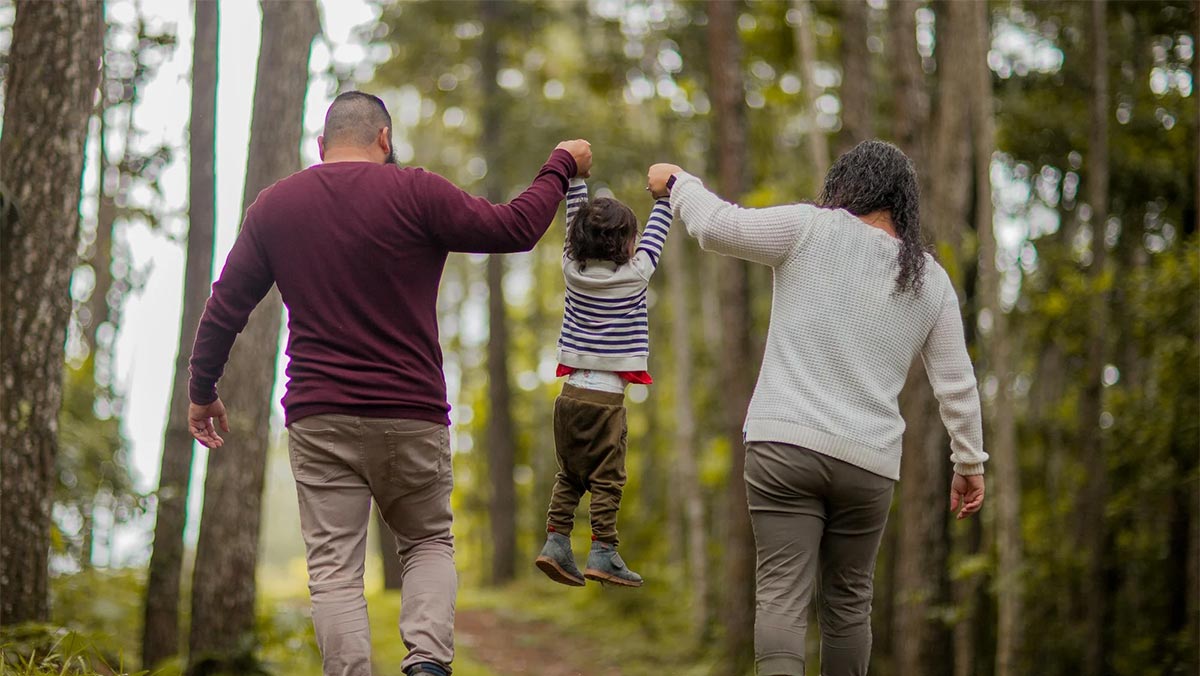 Two parents swing a young child by the hands while walking together through a forest