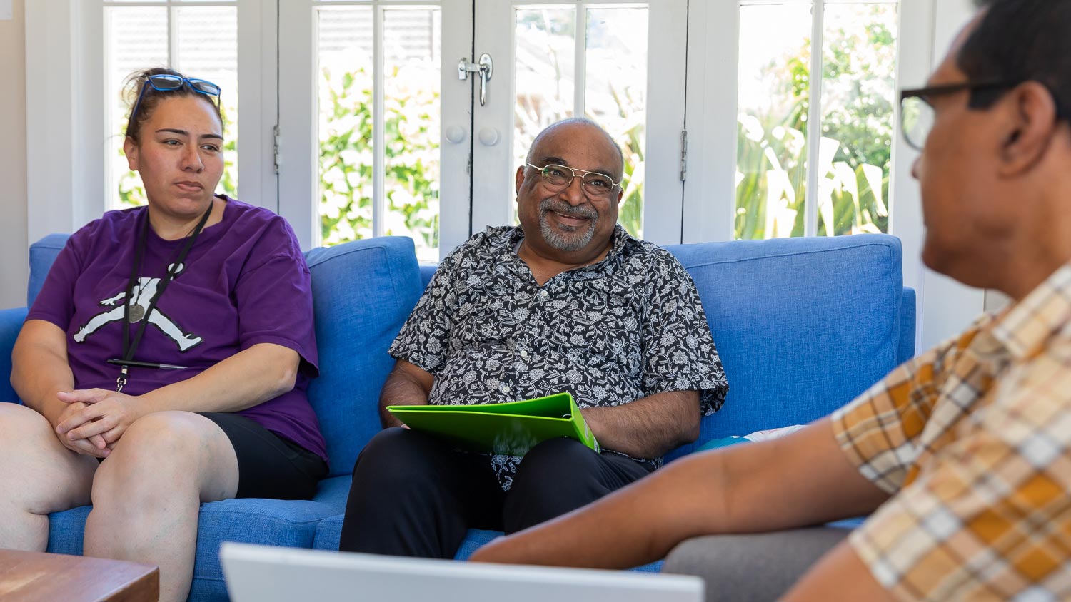 Three people engaged in a discussion indoors, two seated on a blue sofa, one holding a green folder
