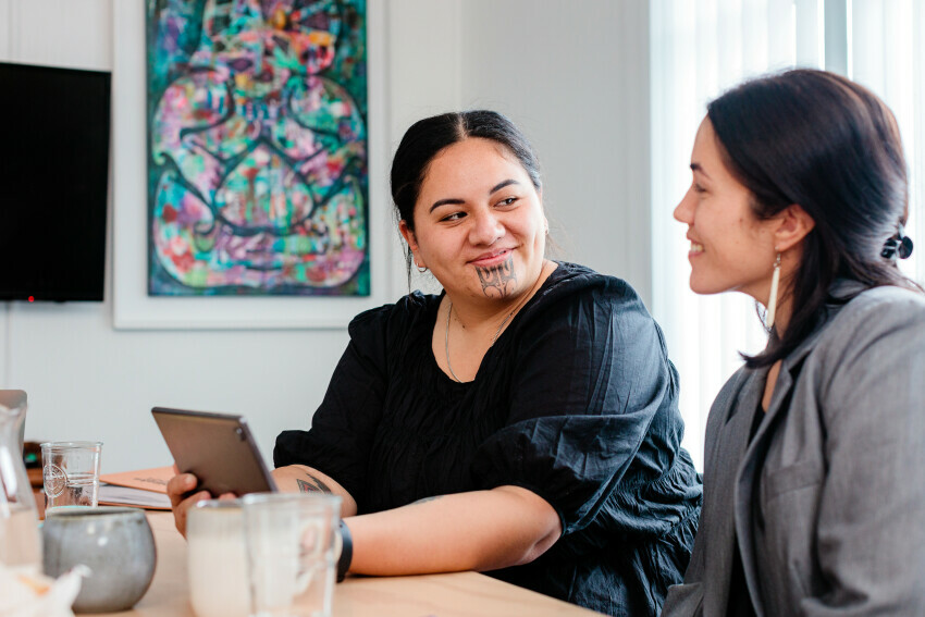 Two women smiling and conversing at a table, one holding a tablet, with colorful artwork on the wall behind them
