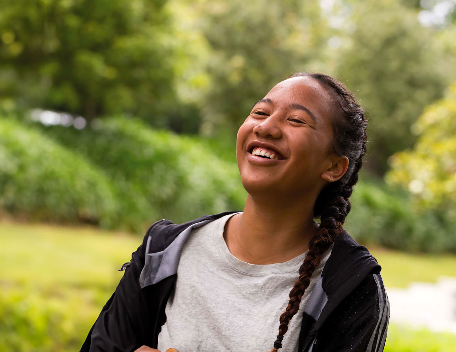 A teenage girl with braided hair laughs joyfully outdoors in a green park setting