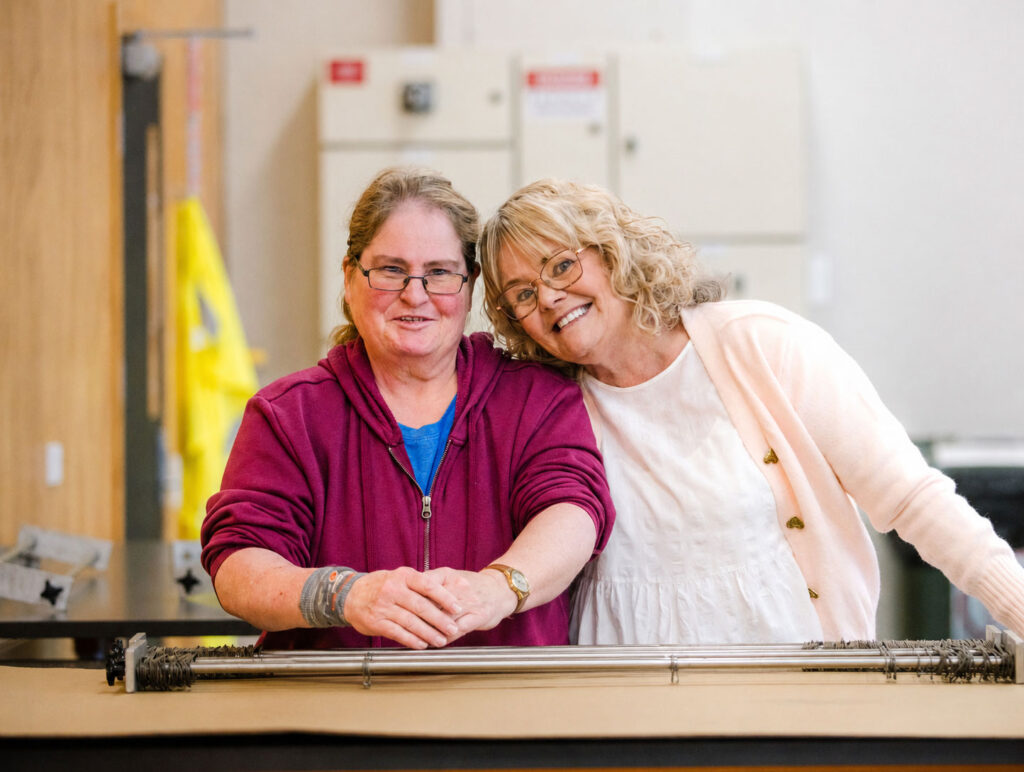 Two smiling women lean together over a workbench in an industrial or workshop setting