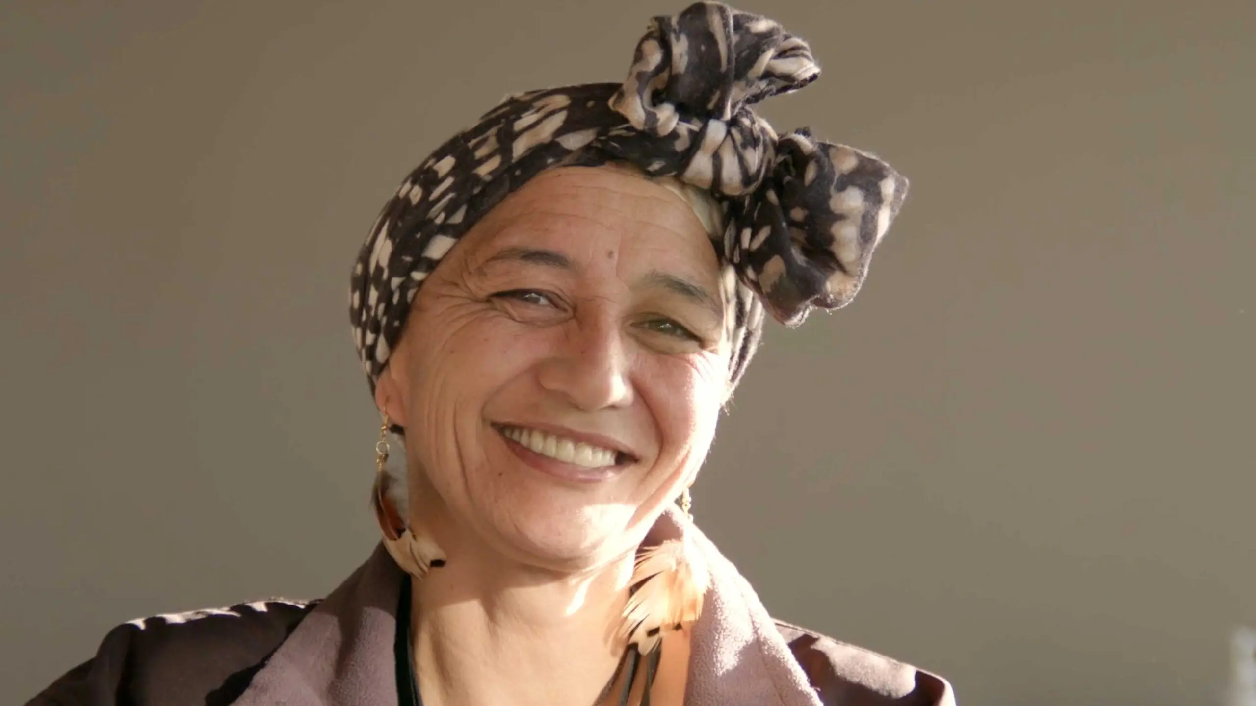 Woman smiling warmly, wearing a patterned head wrap and feather earrings