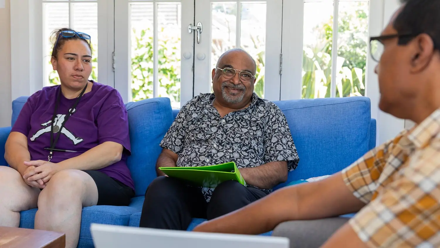 Three people engaged in a discussion indoors, two seated on a blue sofa, one holding a green folder