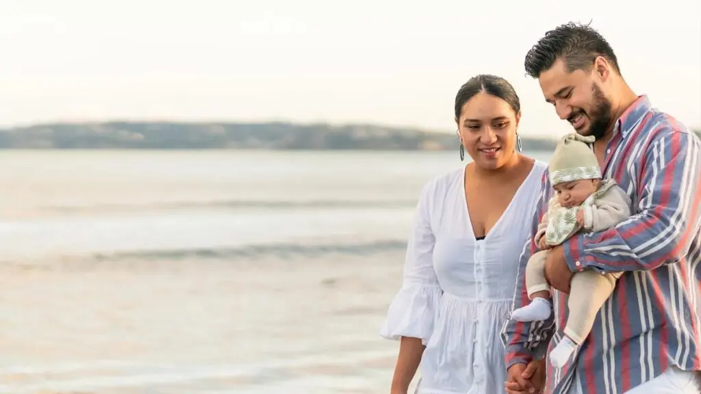 A couple walks along a beach, smiling down at their newborn baby cradled in the man's arms