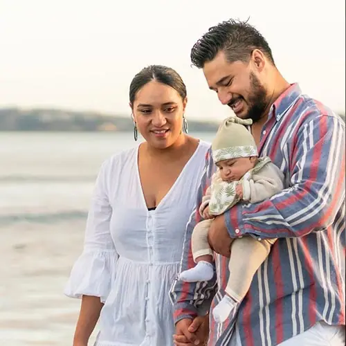 A couple smiling and looking at their newborn baby while walking on a beach