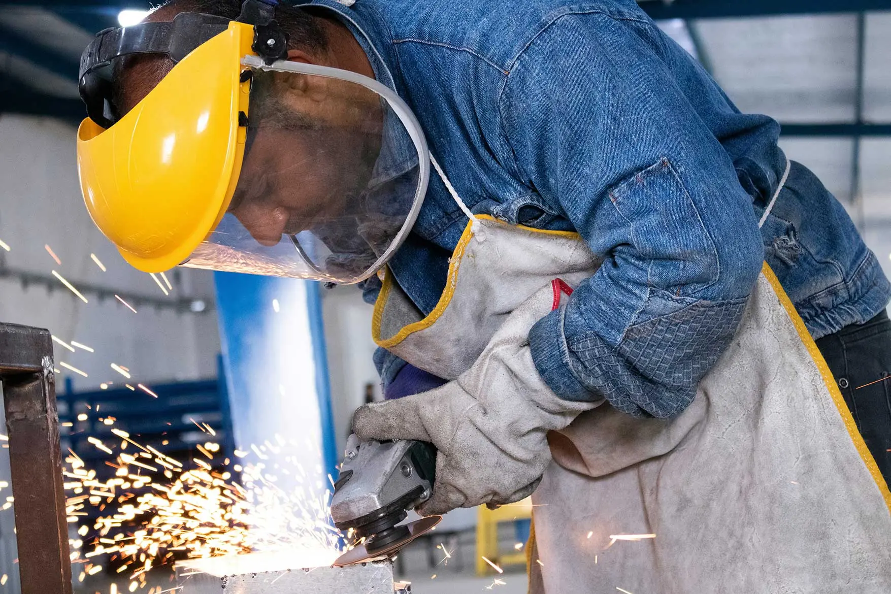 Worker in protective gear and face shield using an angle grinder on metal, sending sparks flying in an industrial facility